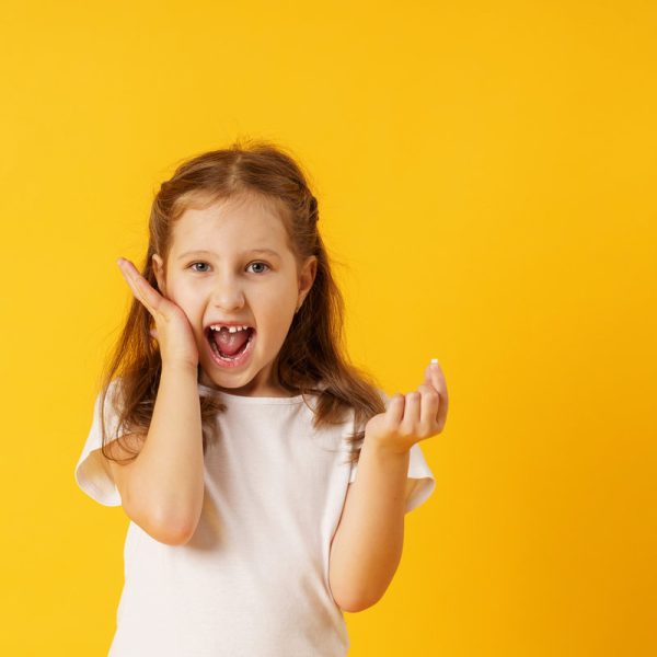 cute smiling preschool girl holds her first fallen baby tooth in her hand while standing on yellow background. growing permanent tooth is in the open mouth. . The concept of hygiene of baby teeth.