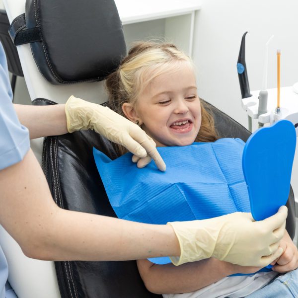 Lovely little girl looking in the mirror at her teeth after teeth surgery in a pediatric stomatology. Happy kid after teeth examination.